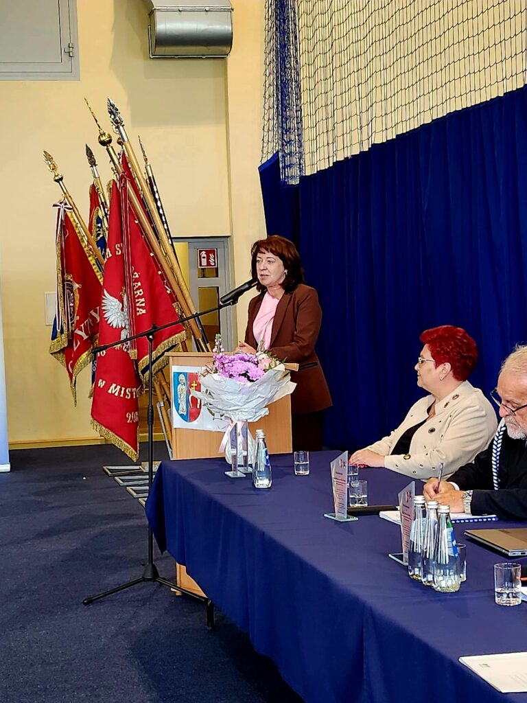Na scenie przemawia kobieta przy mównicy z bukietem kwiatów. W tle wiszą czerwone sztandary na jasnej ścianie. Przy stole z granatowym obrusem siedzą dwie osoby.