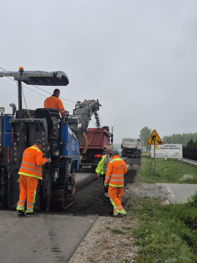 Pracownicy w pomarańczowych strojach naprawiają drogę. Na poboczu znajdują się znaki informujące o utrudnieniach. Szare niebo i zielone pobocze w tle.