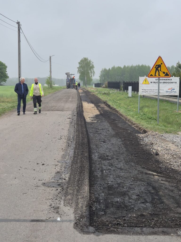Droga w remoncie z pracownikami i sprzętem. Prawa strona drogi jest zdjętym asfaltem. Znaki wskazują na utrudnienia.