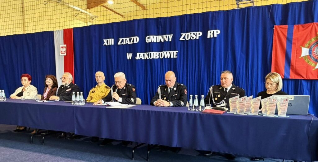 Na zdjęciu widać kilka osób siedzących za stołem na tle niebieskiej kurtyny. W tle widoczne są napisy i flagi. Na stole stoją butelki z wodą i statuetki.