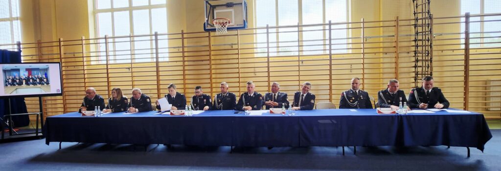 W sali gimnastycznej przy długim stole siedzi grupa strażaków w mundurach. Tło stanowią wysokie okna i drabinki. Obok stołu stoi ekran z grupowym zdjęciem.