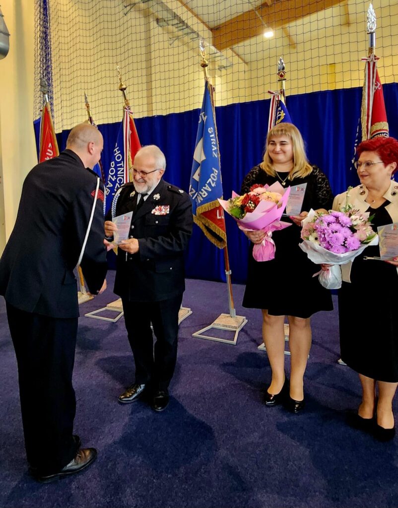 Uroczystość z osobą wręczającą dyplomy i kwiaty. Za nimi stoją flagi na tle niebieskiej kurtyny. Atmosfera oficjalna i elegancka.