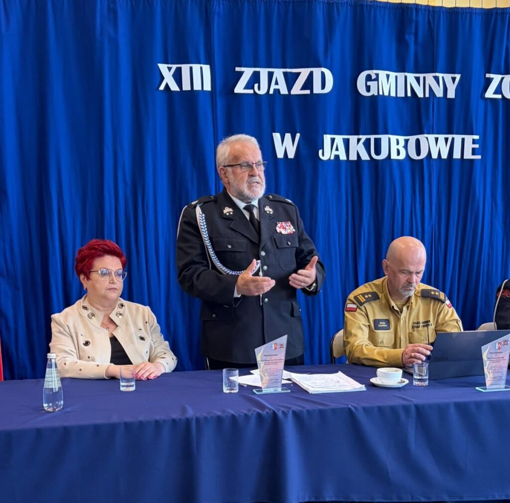 Na zdjęciu są trzy osoby siedzące przy stole. Tło jest niebieskie z białym napisem. Na stole są szklanki i dokumenty.