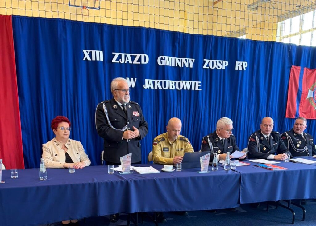 Na zdjęciu siedzi sześć osób przy stole z niebieskim obrusem, tło to niebieska i czerwona tkanina. Mężczyzna w mundurze stoi i przemawia. Napisy na tle wskazują na zjazd w Jakubowie.
