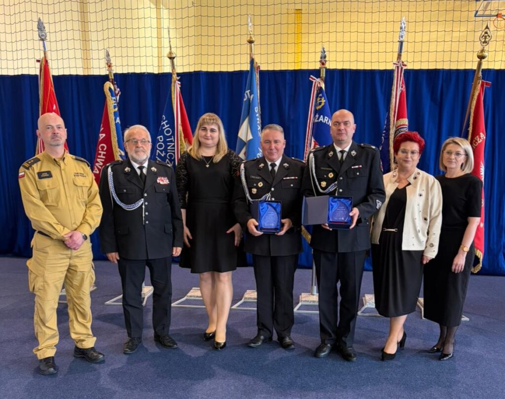 Grupa ludzi w mundurach stoi przed niebieską zasłoną, pośród flag. Wszyscy mają oficjalny strój. Dwa trofea trzymane są na środku.