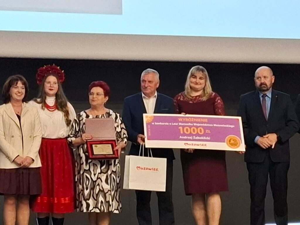 Grupa osób stoi na scenie, trzymając nagrody i czek w kolorze różowym. Jedna osoba w stroju ludowym z czerwoną spódnicą. Tło czarne, u góry białe płótno.