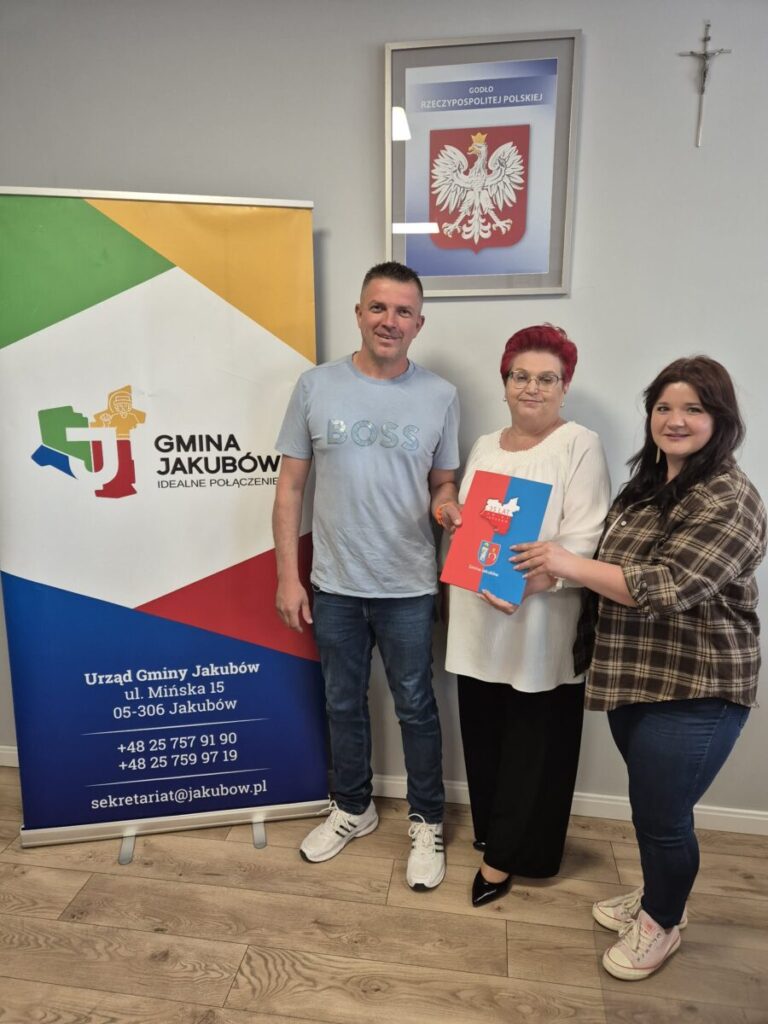 Trzy osoby stoją przed kolorowym banerem gminy Jakubów. Na ścianie wisi godło Polski. Jedna z osób trzyma folder.