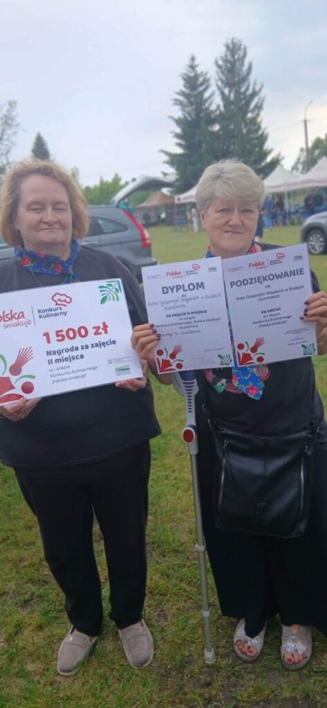 Dwie osoby trzymają dyplomy i czek na 1500 zł na pełnym trawie tle z drzewami i namiotami w tle.