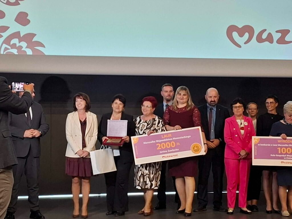 Grupa ludzi stoi na scenie z dużymi czekami. Kobieta w jasnym płaszczu trzyma torbę. Tło jest jasne z logo na górze.