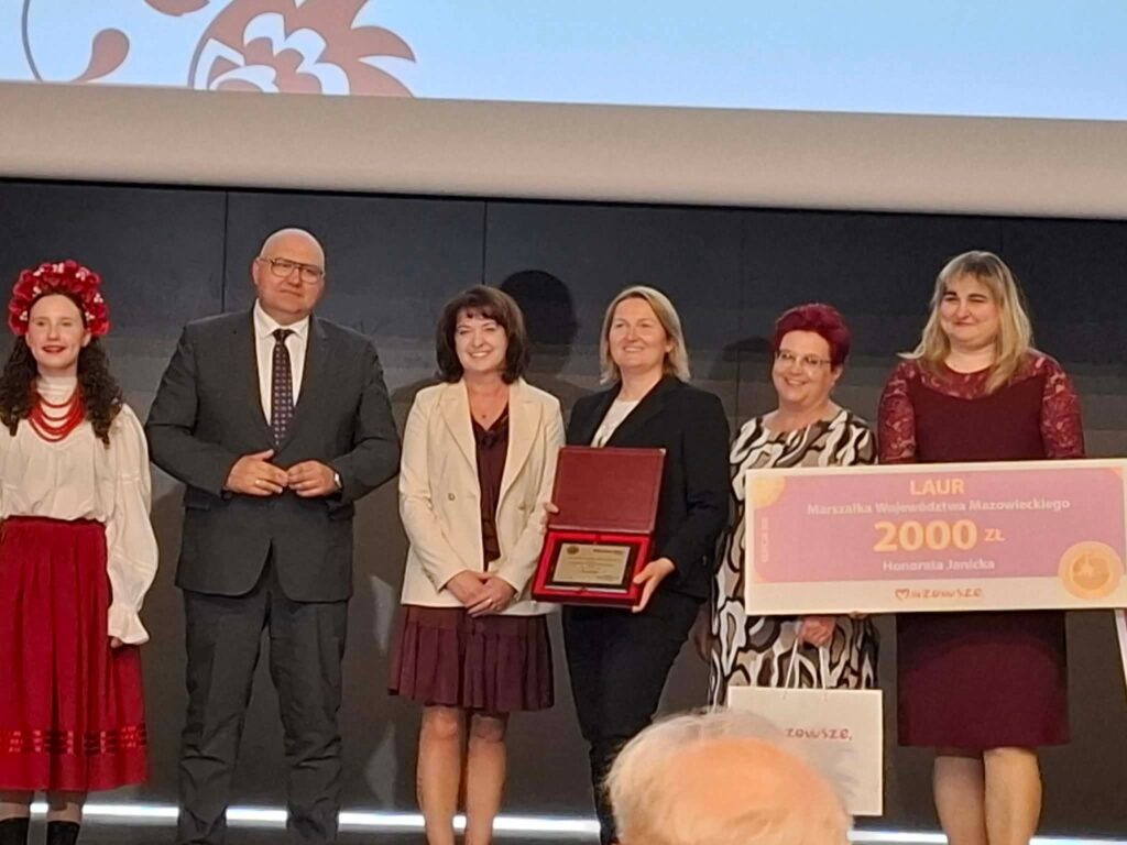 Na zdjęciu sześć osób pozuje przy wręczeniu nagrody. Osoba po lewej ubrana jest w strój ludowy z czerwonym wieńcem. Trzy kobiety trzymają duży czek na 2000 zł, tło jest ciemne.