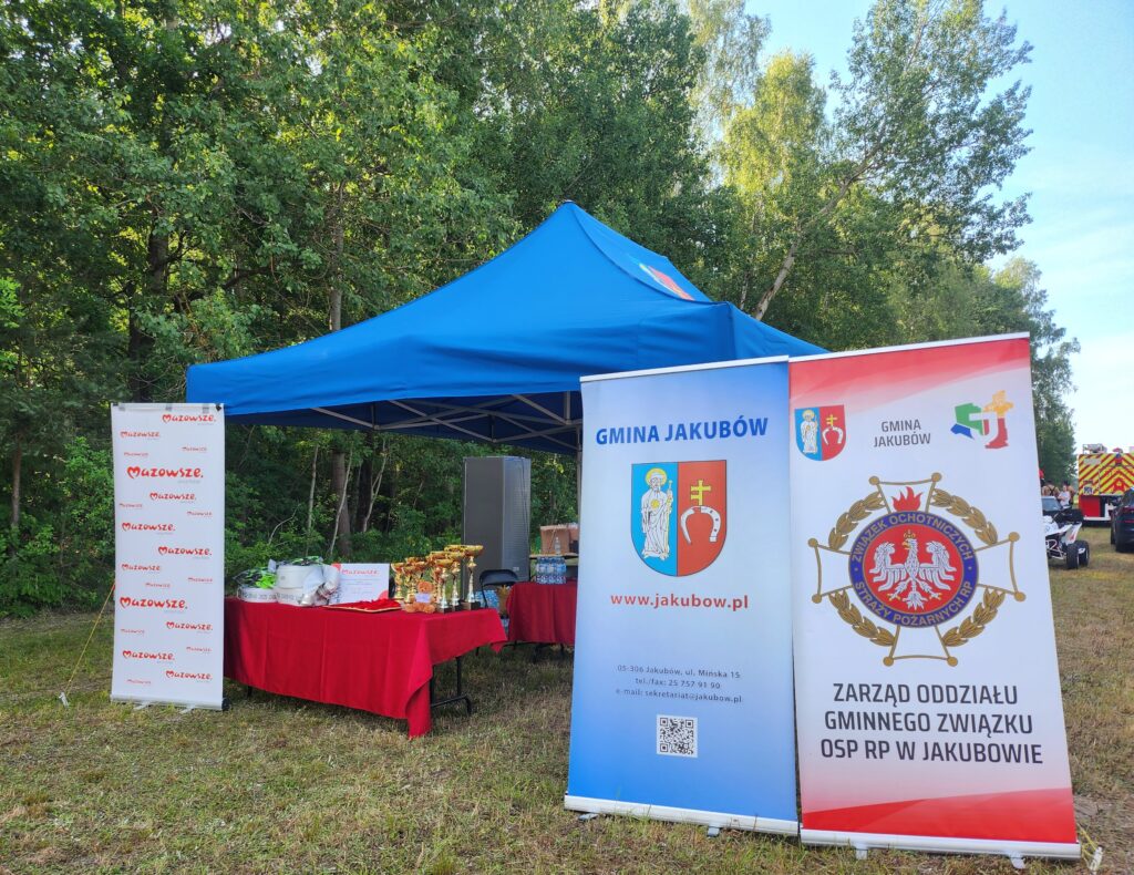 Namiot z niebieskim dachem stoi na tle drzew. Na czerwonym stole ustawione są puchary. Obok widoczne są banery z logo Gminy Jakubów i napisem Mazowsze.