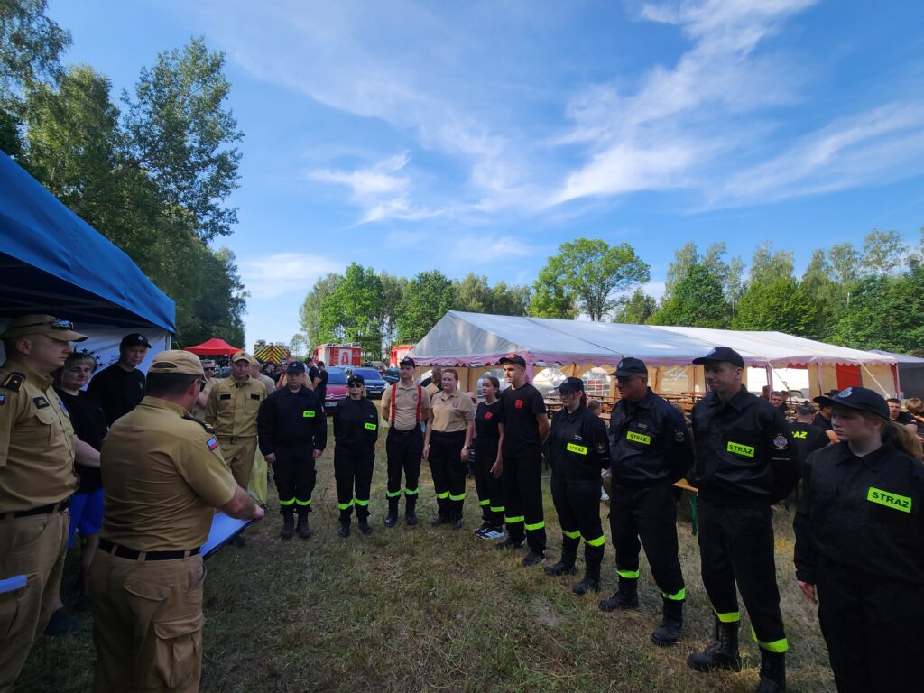 Grupa strażaków w mundurach na zielonym trawniku, tło z namiotami i drzewami, jasne niebo z chmurami.