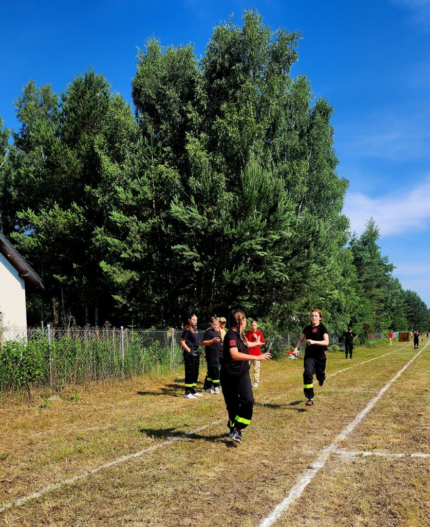 Grupa ludzi biegnie po trawiastej ścieżce w czarnych ubraniach. W tle są zielone drzewa i niebieskie niebo. Obraz przedstawia zawody na świeżym powietrzu.