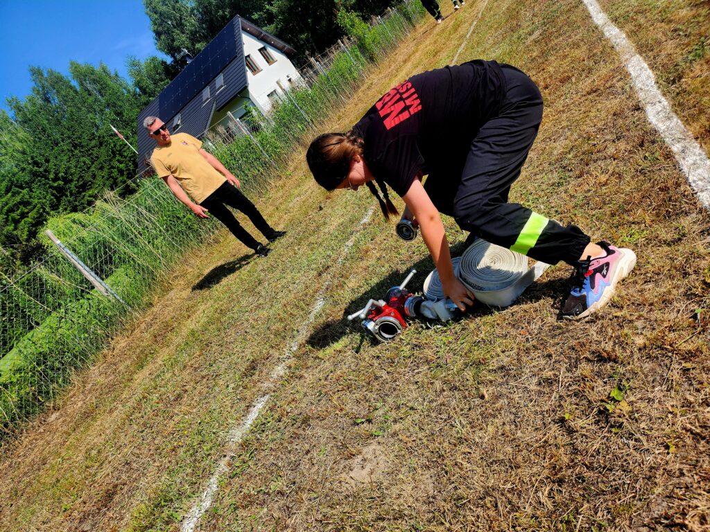 Dziewczyna w czarnym stroju rozwija wąż strażacki na trawie. Obok stoi mężczyzna w beżowej koszulce. W tle jest dom z ciemnym dachem oraz zielone drzewa.