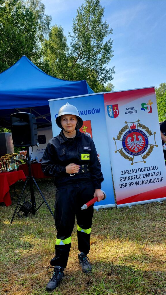 Strażak w stroju i hełmie pozuje z wężem na tle namiotu i banera OSP, w plenerze z drzewami.