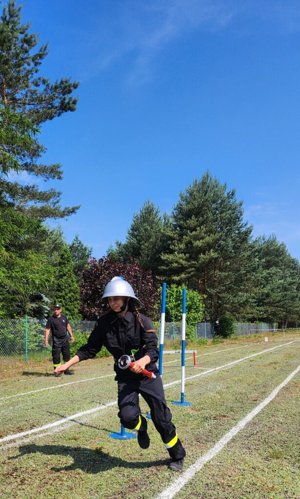 Osoba w stroju strażackim biegnie po torze z przeszkodami. Tło stanowi zielone otoczenie drzew i błękitne niebo. Obok widać metalowe płotki i innego uczestnika.
