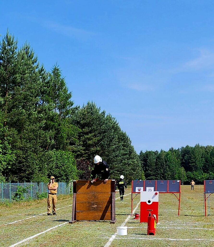 Na zdjęciu strażak w czarnym mundurze przeskakuje przez drewnianą przeszkodę. W tle widać zielone drzewa i niebieskie niebo. Po lewej stoi inna osoba w jasnym ubraniu.