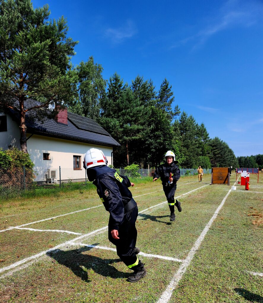 Dwóch strażaków w białych kaskach biegnie po trawiastej ścieżce, na tle niebieskiego nieba, przy białym domu z panelami słonecznymi.