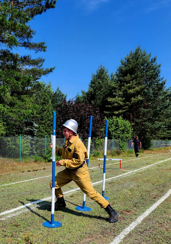 Młoda osoba w żółtym mundurze biegnie między niebieskimi słupkami na zielonej trawie. W tle widać ogrodzenie i drzewa. Niebieskie niebo.