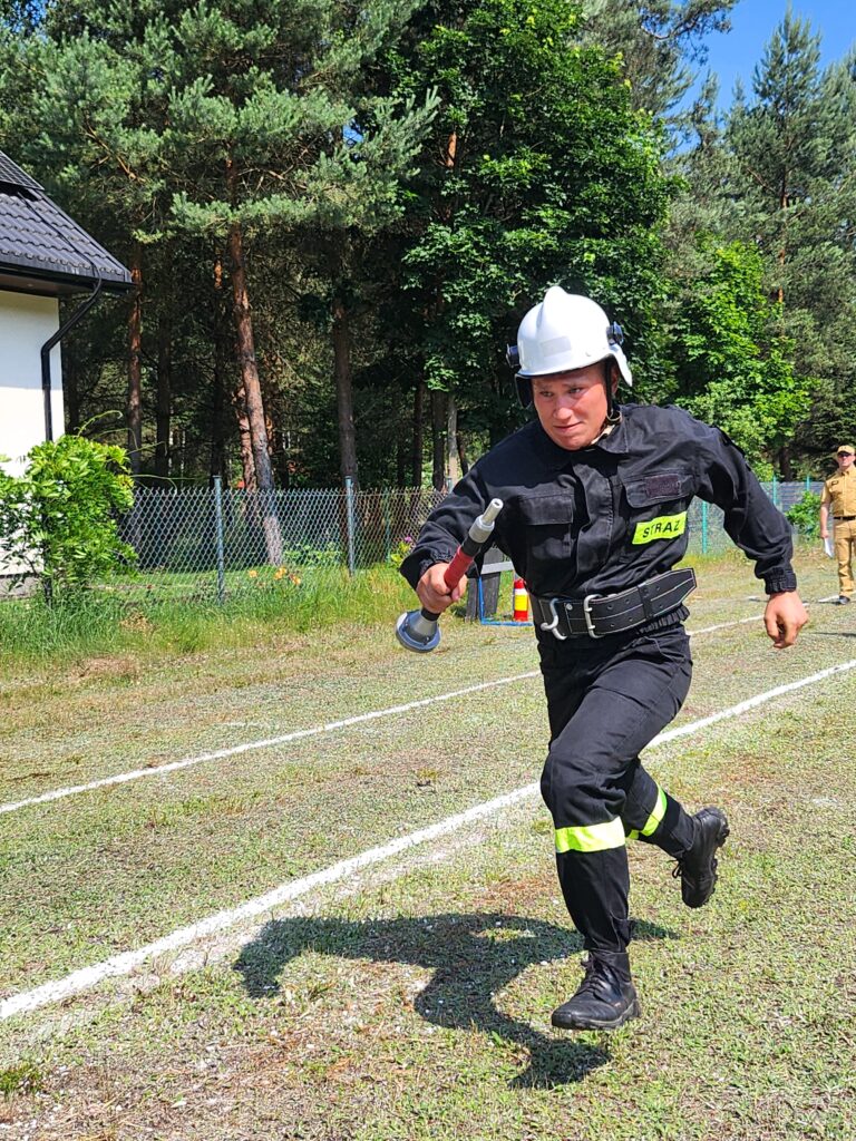 Strażak w czarnym uniformie i białym kasku biegnie po trawie. W tle las i fragment budynku z ciemnym dachem. Dzień słoneczny.