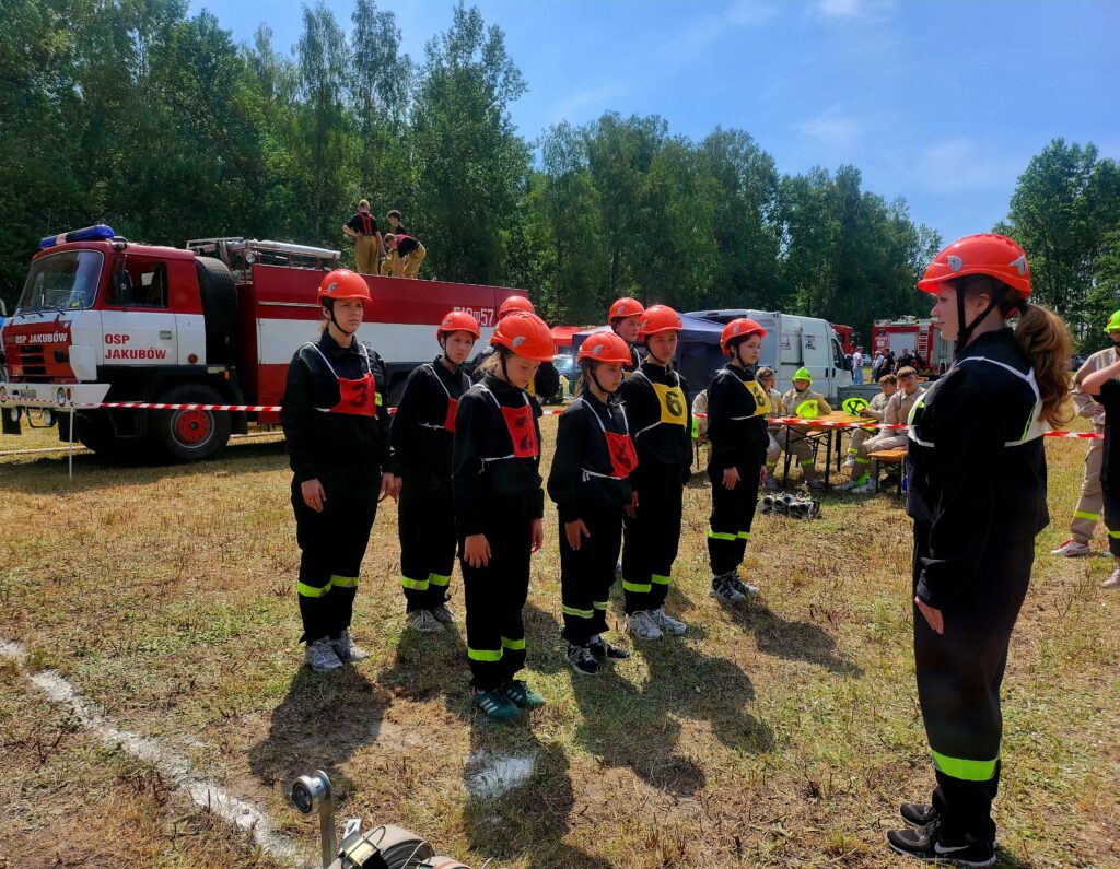 Grupa młodych strażaków w czarnych mundurach i czerwonych kaskach stoi na trawie. W tle widać czerwony wóz strażacki i drzewa. Niebo jest jasnoniebieskie, a teren ogrodzony taśmą.