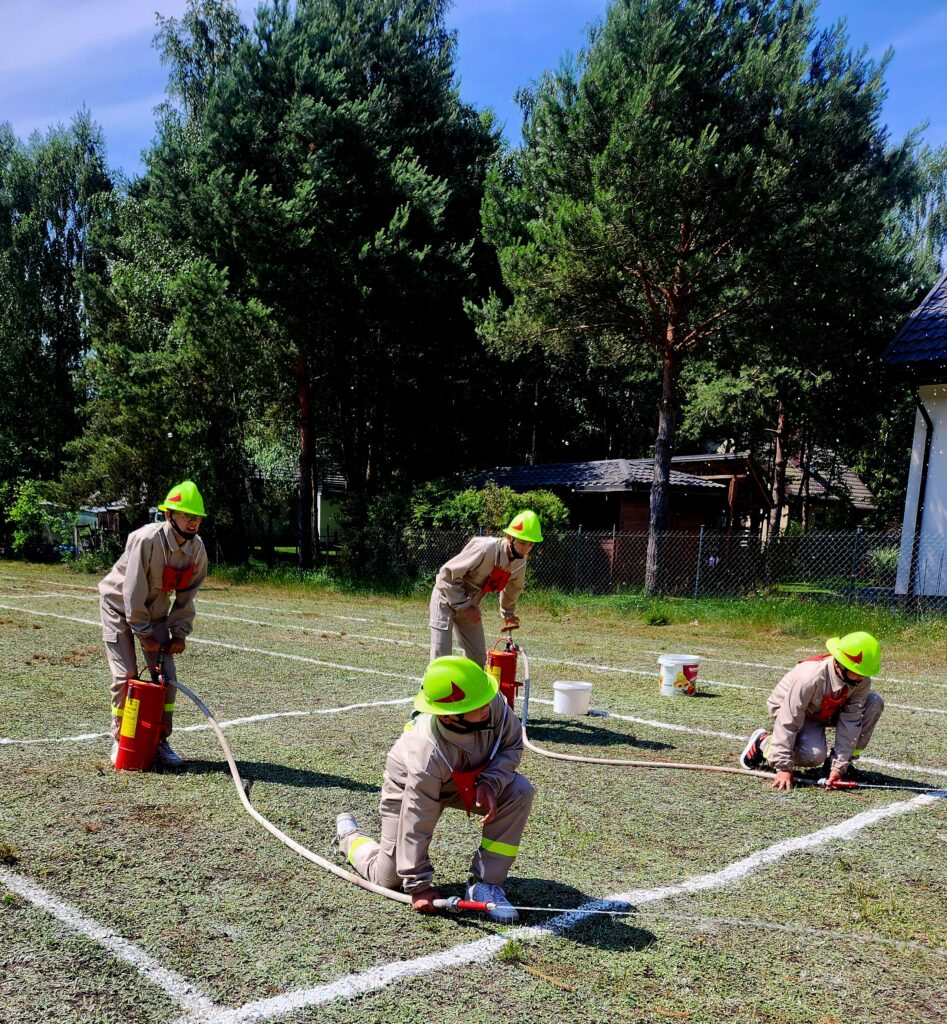 Grupa osób w kaskach neonowych symuluje działania strażackie na trawniku. Ubrani są w jasne kombinezony, z czerwonymi akcentami. W tle bujna zieleń drzew i drewniana szopa.