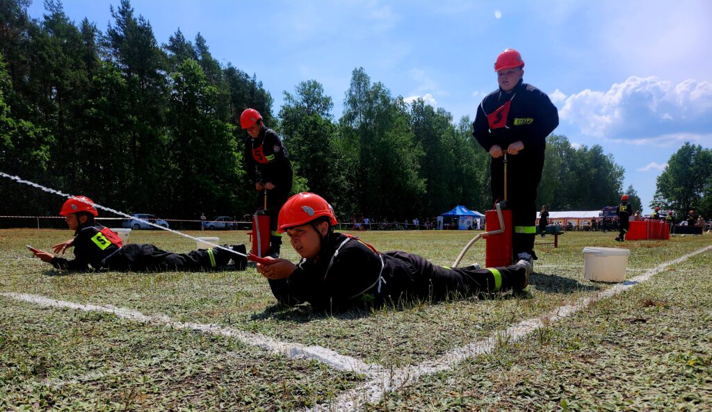 Dzieci w kaskach ćwiczą strażackie zadania na tle zielonego lasu. Niebieskie niebo, biały namiot w tle. Jeden z nich leży na ziemi z wężem do wody.
