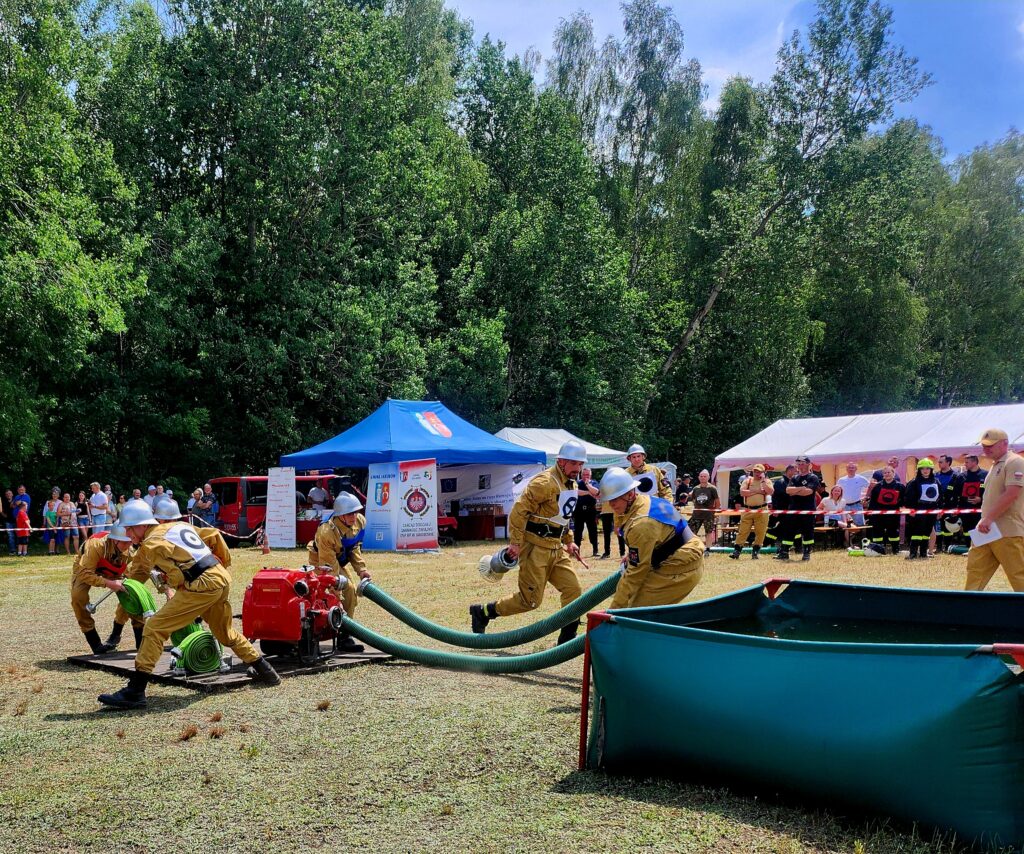 Grupa strażaków w żółtych mundurach z hełmami ćwiczy, używając węży przy zielonym zbiorniku. W tle są dwa namioty: niebieski i biały oraz wielu obserwatorów. Sceneria otoczona jest gęstym zielonym lasem