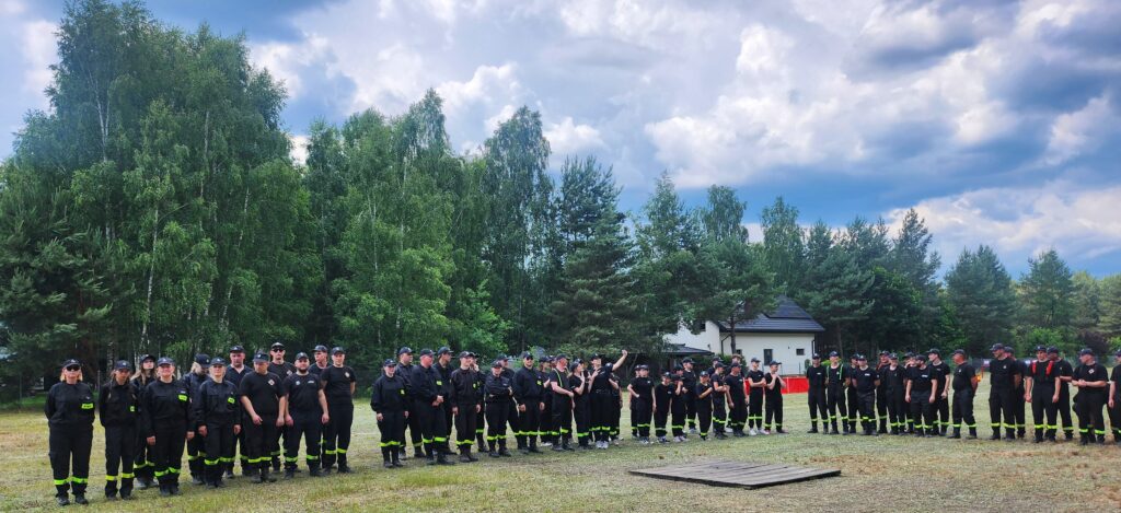Grupa osób w czarnych mundurach stoi na trawie. W tle zielone drzewa i niebo z chmurami. Bocznie widoczny biały budynek.