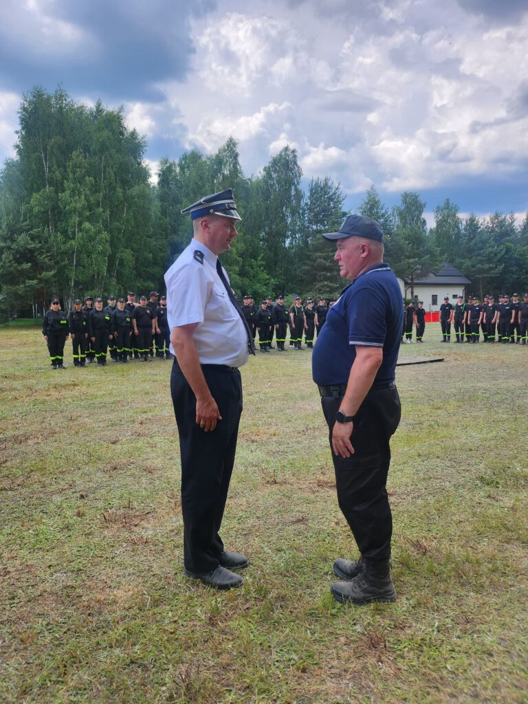 Dwaj mężczyźni stoją naprzeciw siebie na trawie. Jeden ma białą koszulę z ciemnym krawatem, drugi granatową polo. W tle las i grupa ludzi w czarnych mundurach.