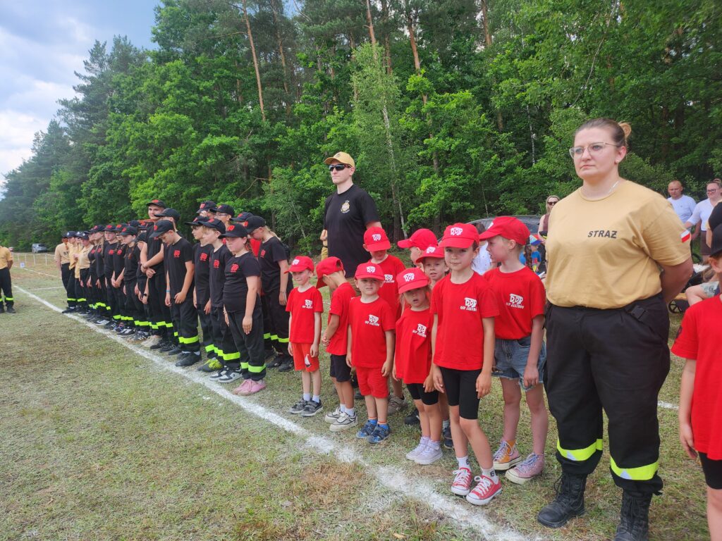 Grupa dzieci i młodzieży w czerwonych i czarnych strojach stoi w szeregu na trawie. Tło stanowi zieleń drzew. Dorosły w beżowej koszulce z napisem STRAŻ stoi na przodzie.