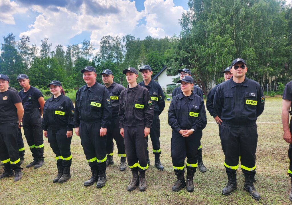 Grupa osób w czarnych mundurach strażackich stoi na trawie. Na ramionach mają żółte opaski z napisem STRAŻ. W tle zielone drzewa i niebo z chmurami.