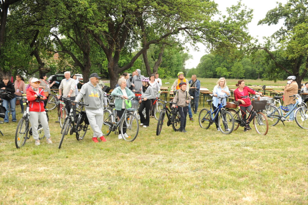 Na zdjęciu znajduje się grupa osób z rowerami na zielonej trawie. W tle widać drzewa i ustawione stoły piknikowe. Uczestnicy ubrani są w luźne, sportowe ubrania.
