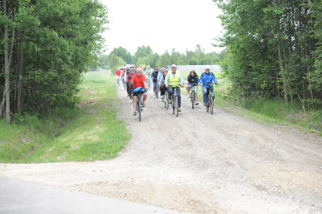 Grupa rowerzystów jedzie leśną drogą, uśmiechnięci, ubrani kolorowo. Dookoła zielone drzewa i trawa, niebo jasne. Tło naturalne, spokój.