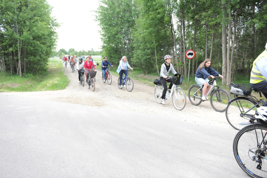 Grupa rowerzystów jedzie szutrową drogą przez zalesiony teren. Ubrania są kolorowe, dominują zieleń i błękit. W tle widać las oraz znak drogowy.