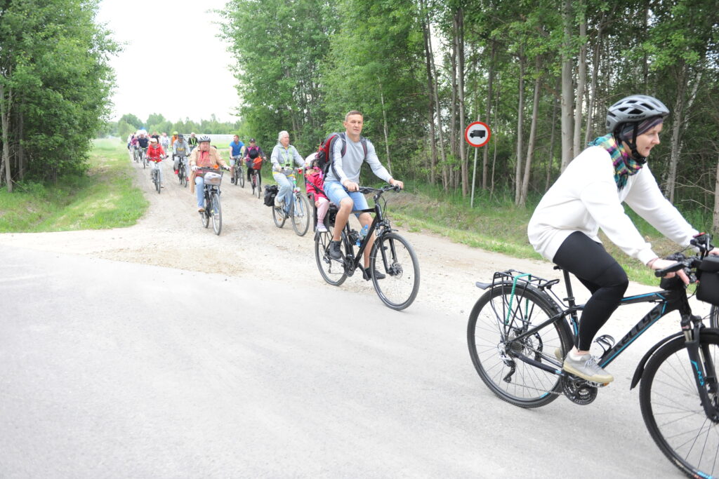 Grupa rowerzystów jedzie po żwirowej drodze otoczonej zielonym lasem. Osoby mają kolorowe ubrania. W tle znak zakazu wjazdu dla samochodów.