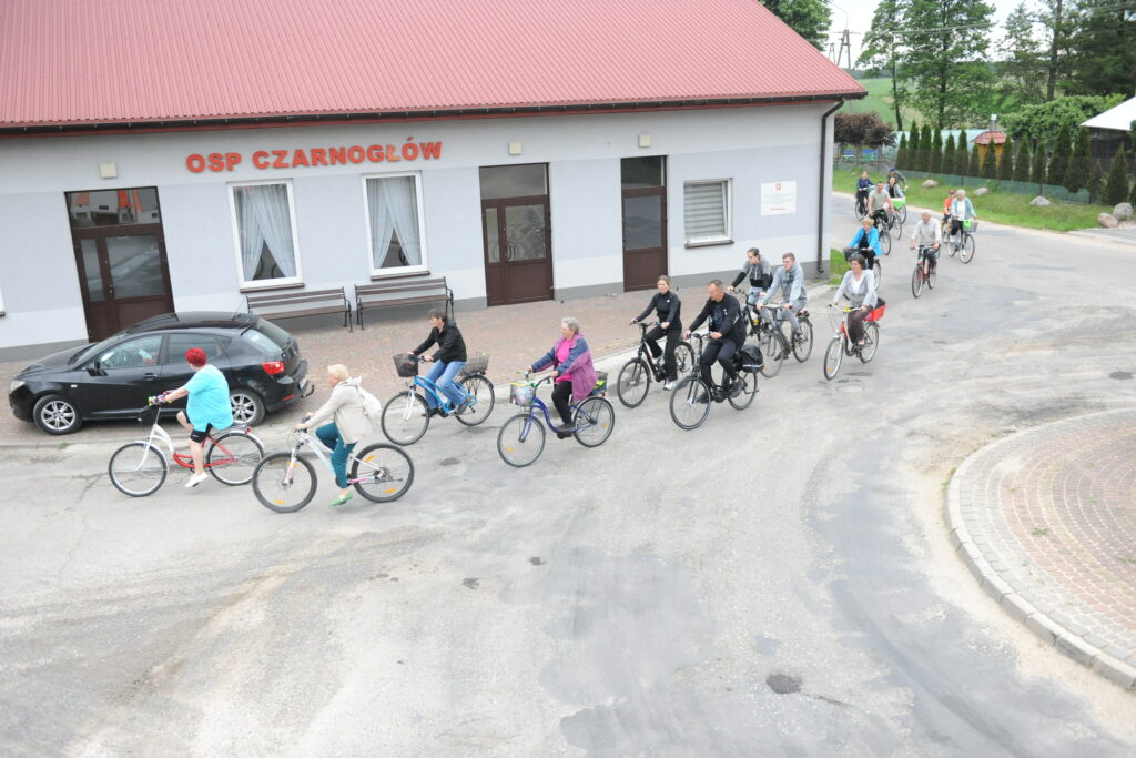 Grupa rowerzystów jedzie ulicą przed szarym budynkiem z czerwonym dachem. Na budynku napis OSP Czarnogłów. Tło to zielone drzewa i chodnik.