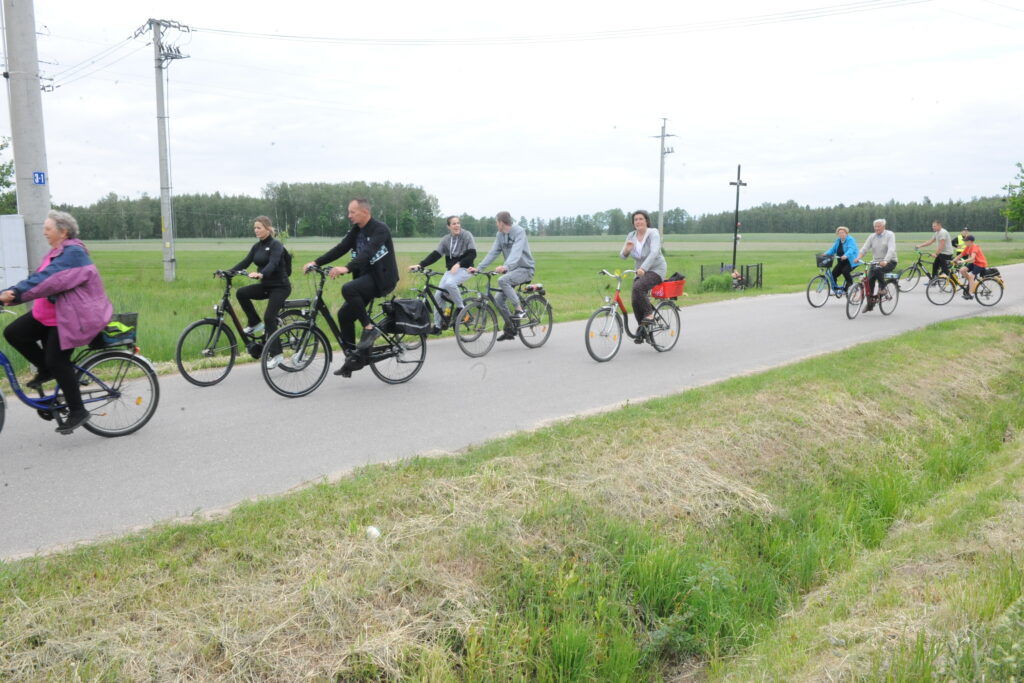 Grupa ludzi jedzie na rowerach po wiejskiej drodze. Tło stanowią zielone pola i las. Pogoda jest pochmurna, a ludzie noszą kolorowe ubrania.