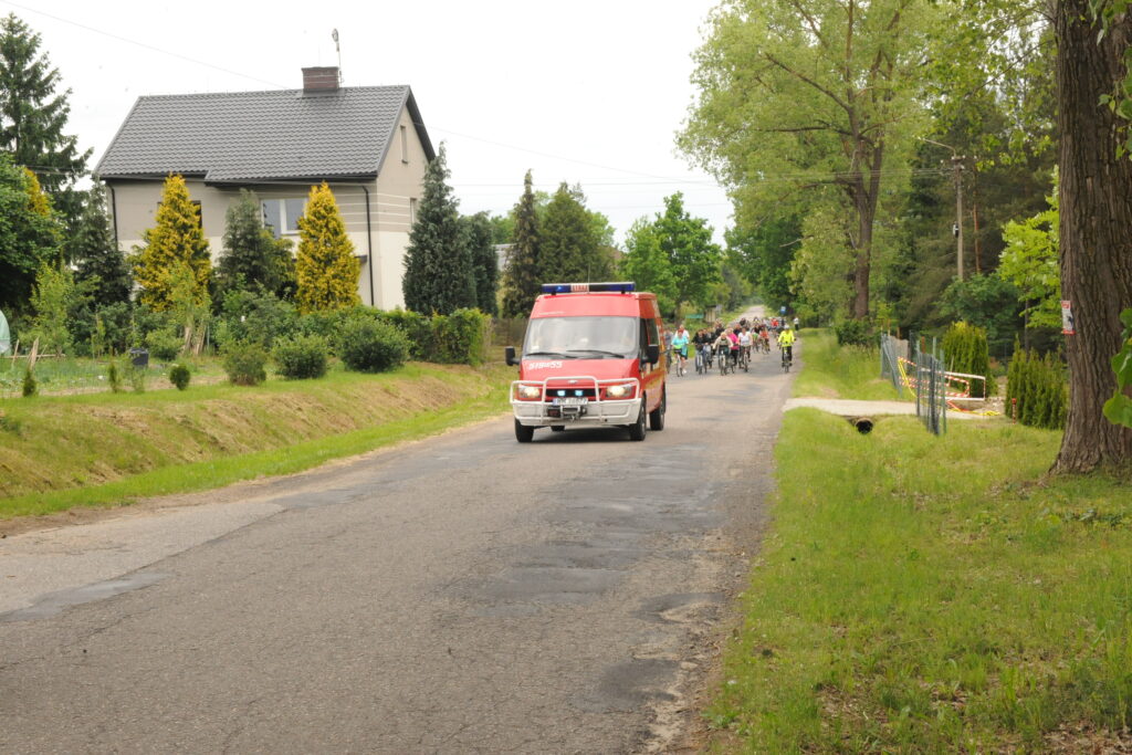 Czerwona karetka jedzie wąską wiejską drogą, otoczoną zielenią i drzewami. W tle grupa rowerzystów. Po lewej, dom wśród krzewów.