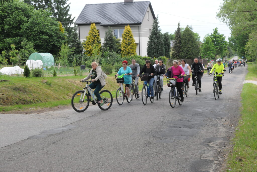 Grupa rowerzystów jedzie asfaltową drogą w zielonej okolicy. W tle dom jednorodzinny i ogródki. Uczestnicy ubrani kolorowo.