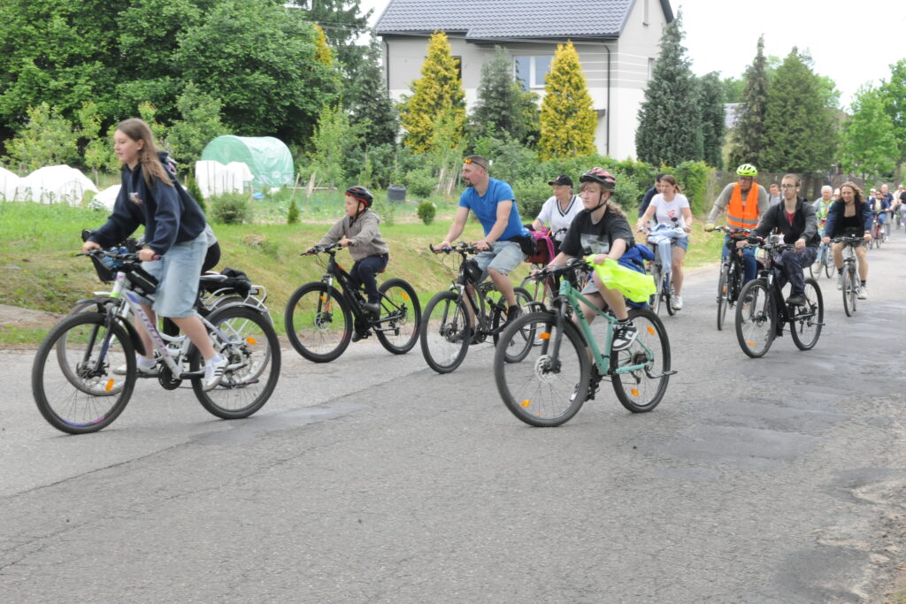 Grupa rowerzystów jedzie po asfaltowej drodze. Ubrane są w różnorodne stroje, a w tle widać zieleń i dom. Niebo jest jasne, co sugeruje słoneczny dzień.
