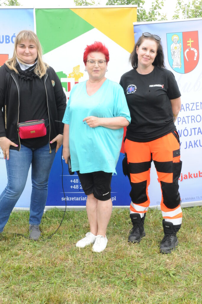 Trzy osoby stoją na trawie przed kolorowymi banerami. Jedna ma czerwone włosy, druga okulary przeciwsłoneczne na głowie. Osoby noszą niebieską, czarną i pomarańczową odzież.