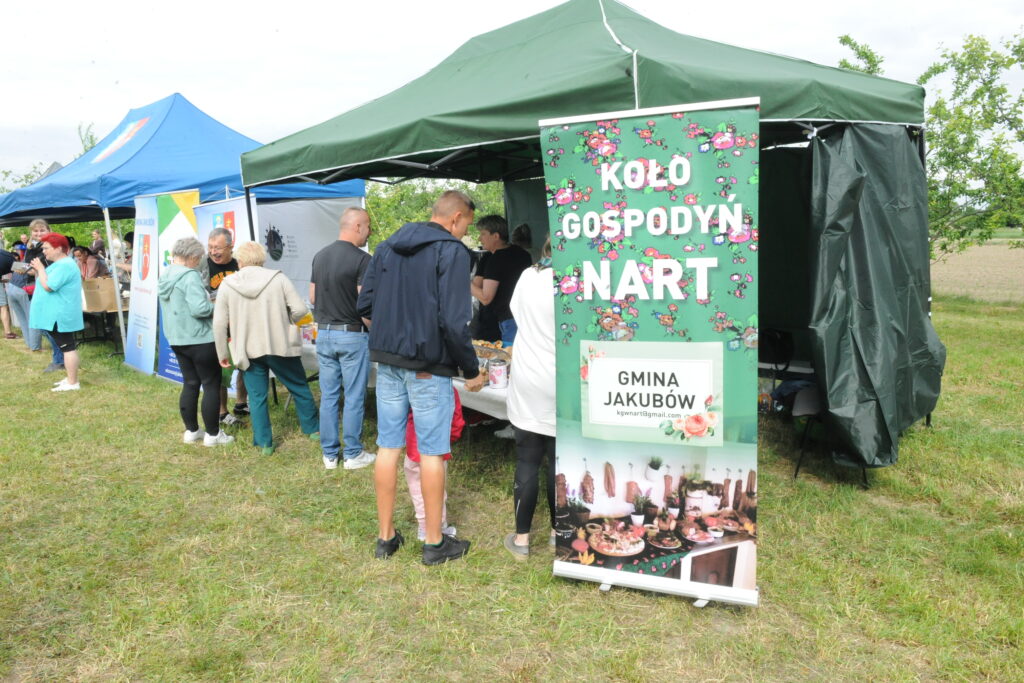 Na zdjęciu widoczny jest zielony namiot z napisem Koło Gospodyń Nart. Obok niebieski namiot na tle zielonej trawy. Ludzie stoją w kolejce przy stoiskach.
