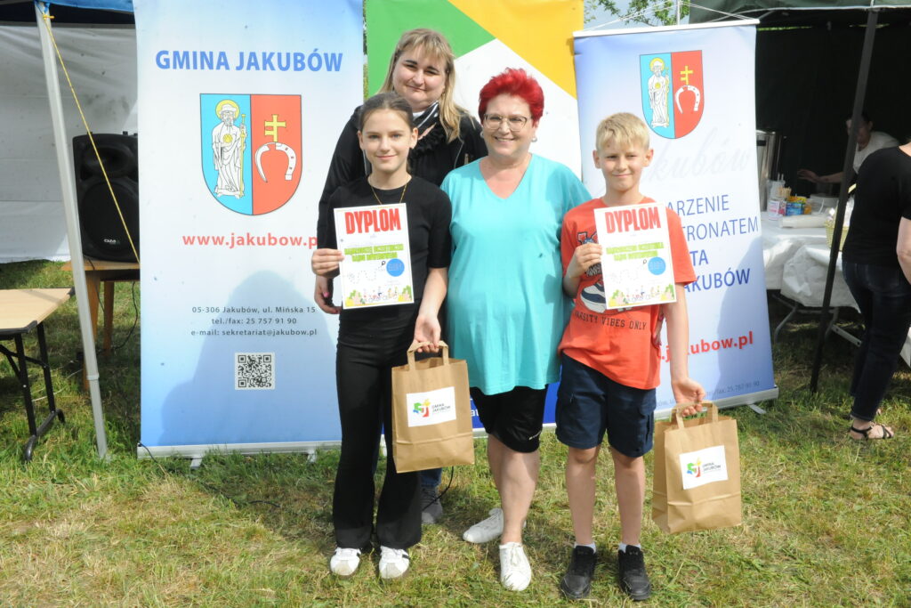 Na zdjęciu czwórka osób trzymających dyplomy i torby. Tło z plakatami i zieloną trawą. Kolory jasne, przewaga niebieskiego i zielonego.
