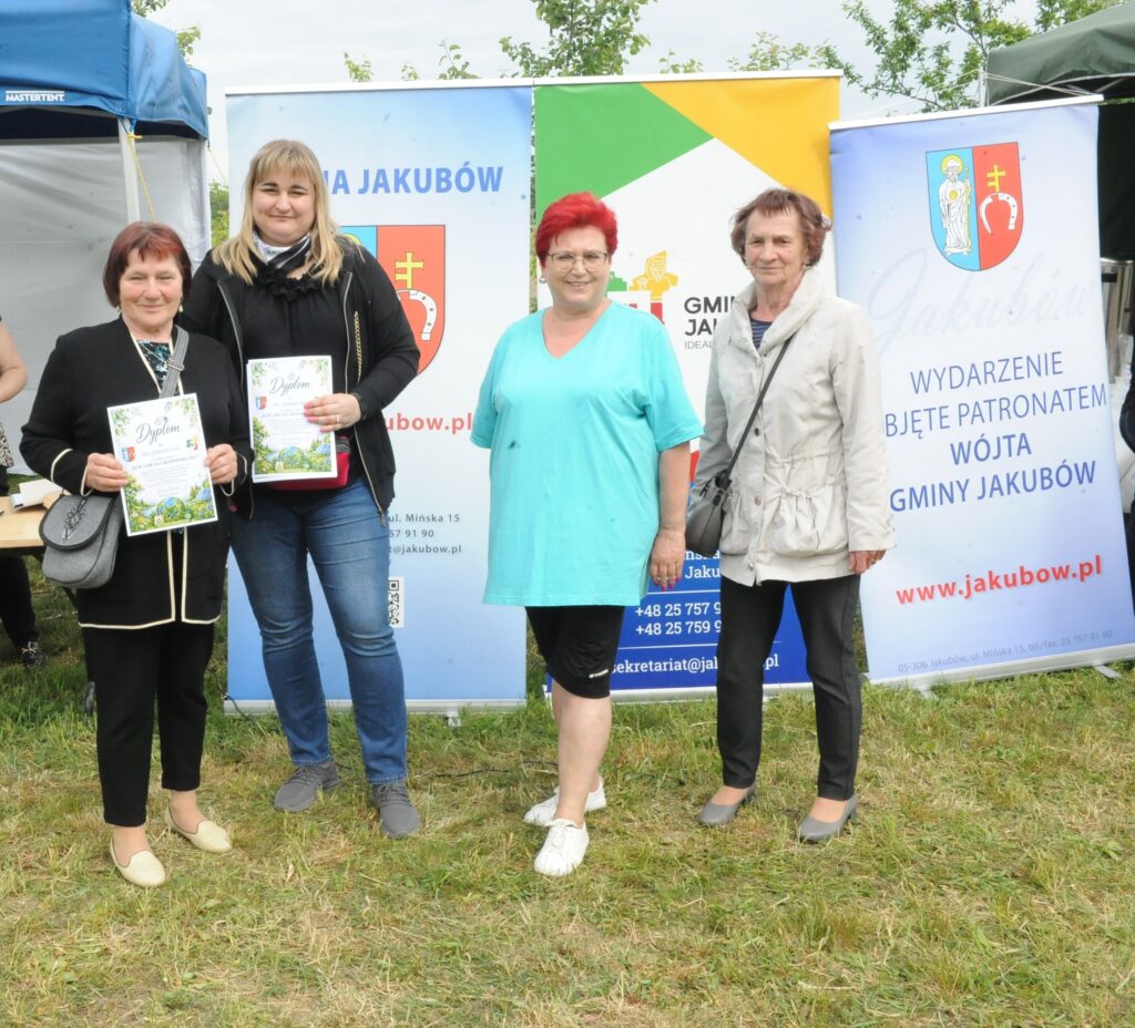 Na zdjęciu cztery osoby stojące na trawie przed stoiskami z banerami w tle. Dwie osoby trzymają dyplomy, a kolorystyka zdobi niebieskie i zielone odcienie. Tło zawiera informacje dotyczące gminy Jakubów.