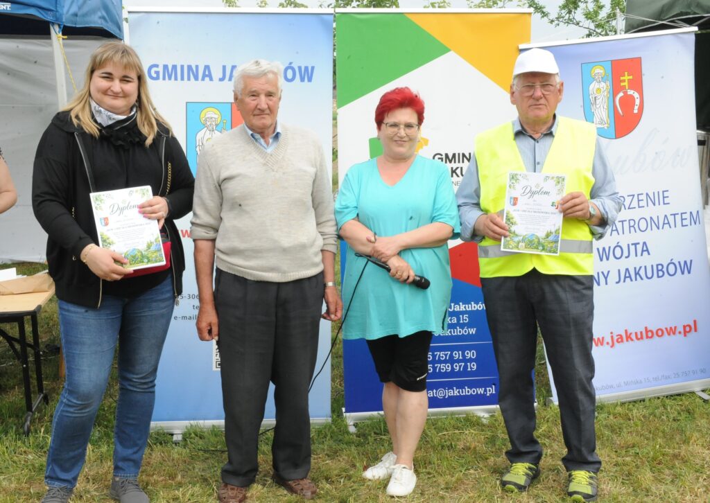 Na zdjęciu są cztery osoby stojące przed kolorowymi banerami. Dwóch z nich trzyma dyplomy, jedna osoba ma czerwoną koszulę i mikrofon. Tło to jasnozielony trawnik z tekstami na banerach.