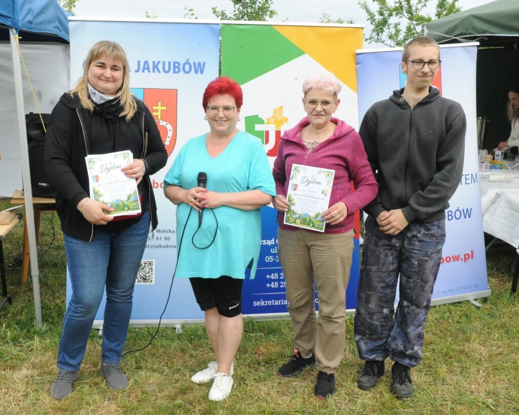 Cztery osoby stoją na trawie z dyplomami; tło z plakatami pełne jasnych barw. Jedna osoba trzyma mikrofon.