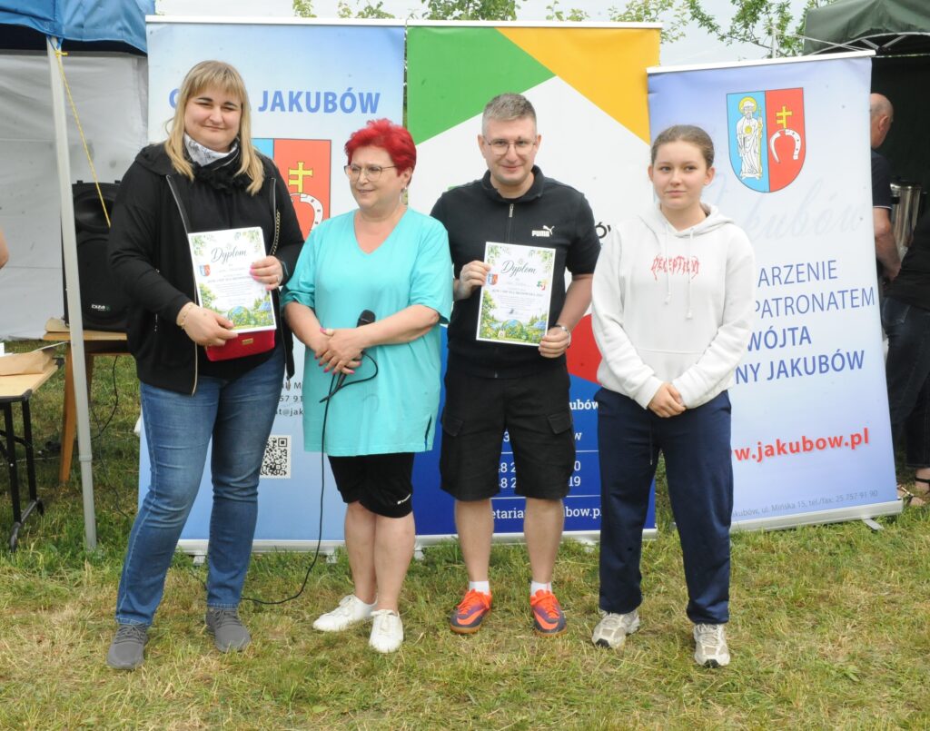 Na zdjęciu widać cztery osoby stojące przed kolorowymi banerami promocyjnymi. Osoby trzymają dyplomy, ubrani są w codzienne stroje. Tło to zielona trawa i niebo, sugerujące plenerową scenerię.