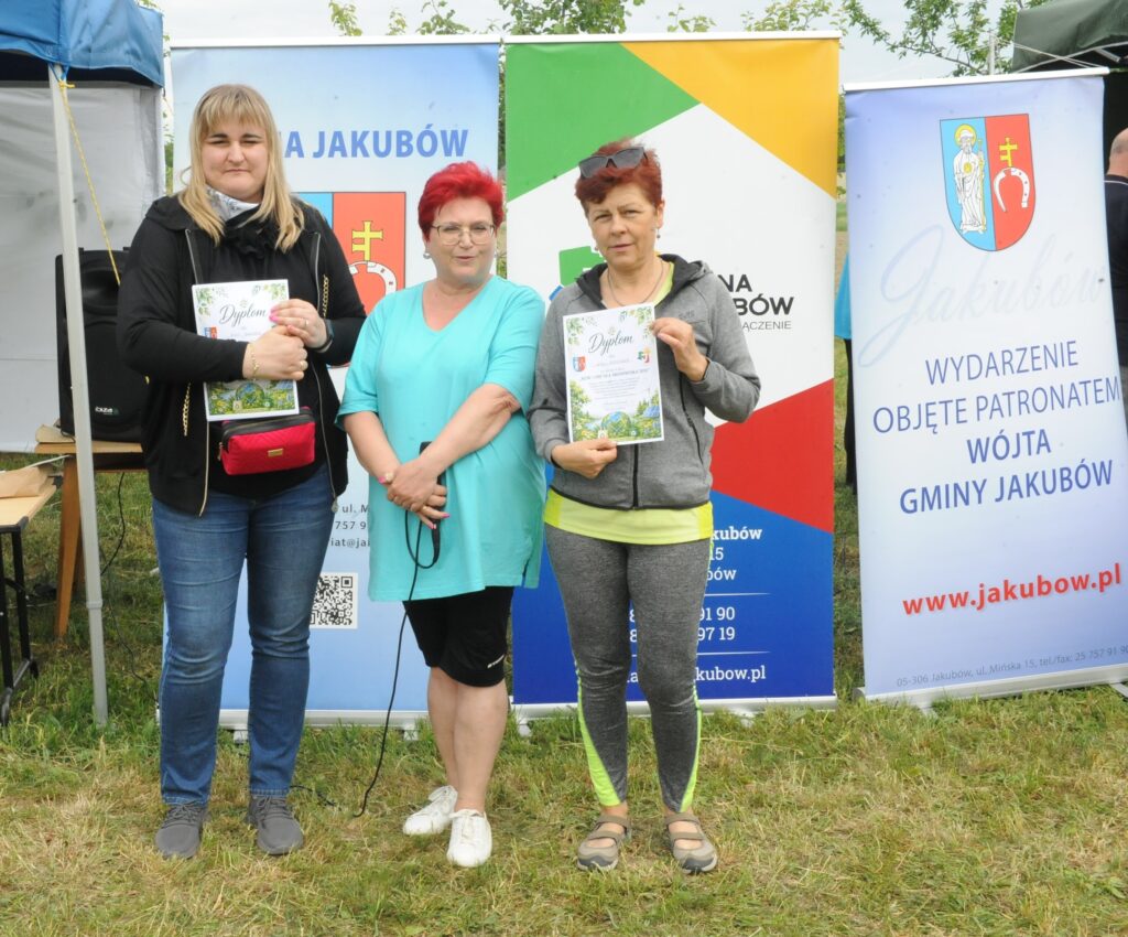 Trzy kobiety stoją na tle kolorowych banerów, trzymając dyplomy. Trawa pod stopami kontrastuje z jasnym, pochmurnym niebem. Środkowa kobieta ubrana w turkusową bluzkę.