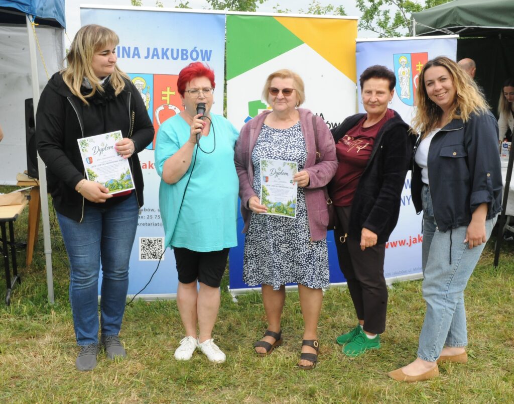 Na zdjęciu pięć osób stoi na trawie, trzymając dyplomy. Tło to banery promocyjne z kolorami zielonym, niebieskim i białym. Osoby wyglądają na zadowolone, a pogoda jest słoneczna.